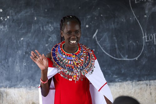 Girl in Maasai dress stands in front of a blackboard in a classroom, smiling as she explains something