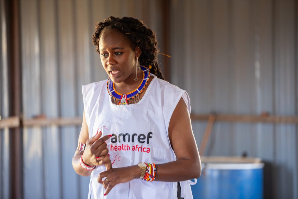 Cynthia stands, speaking passionately. She has an audience that isn't pictured. She wears an Amref tabard.