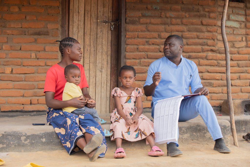 A mother sits outside on a step with her young son on her lap and her daughter beside her. To their right, a health worker in blue scrubs and a large notebook sits, speaking to the mother.