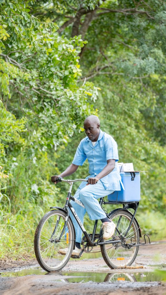 A healthcare worker cycles around a puddle. He wears blue scrubs and has a blue cool box strapped to his bike.