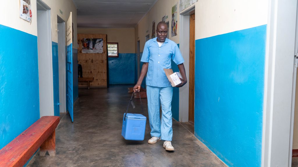 A health worker stands in a clinic corridor wearing blue scrubs. He carries a large blue cool box in one hand boxes in the other.