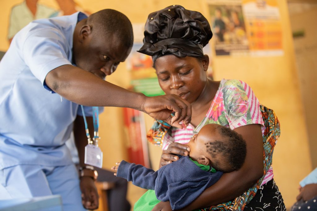A healthcare worker leans over a mother holding a young toddler in her arms to administer a vaccine orally