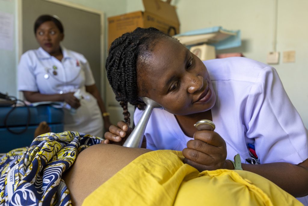 A midwife uses a horn to listen to a baby's heartbeat in utero. She is in a hospital room and a nurse waits in the background.