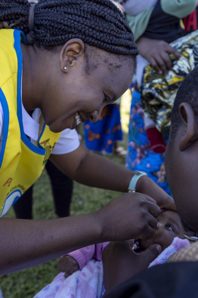 A close up of a nurse smiling at a baby, in her mother's arms, as she administers a vaccine orally. They are outside and there is a queue of people waiting.