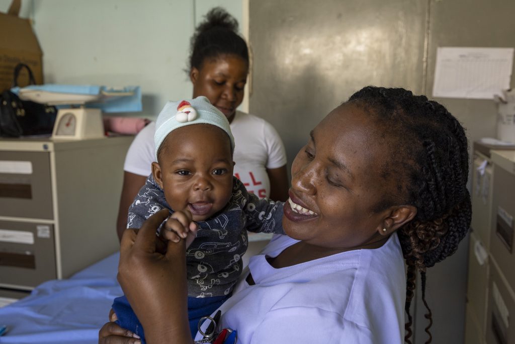 A baby is being held in a nurse's arms. He is holding onto her hand and reaches out to the camera. His mother is in the background. Everyone is smiling.