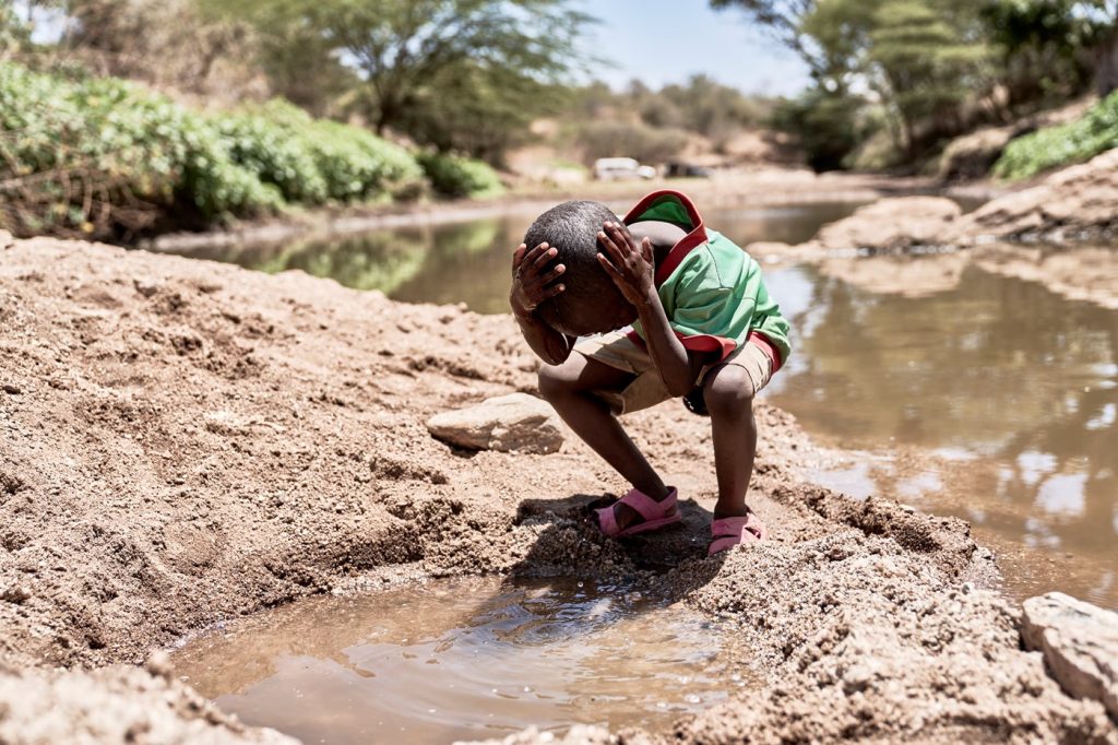 A young boy leans forward with his head in his hands in front of a dirty puddle of water. He is at a small river and there are trees and he is standing in soil.