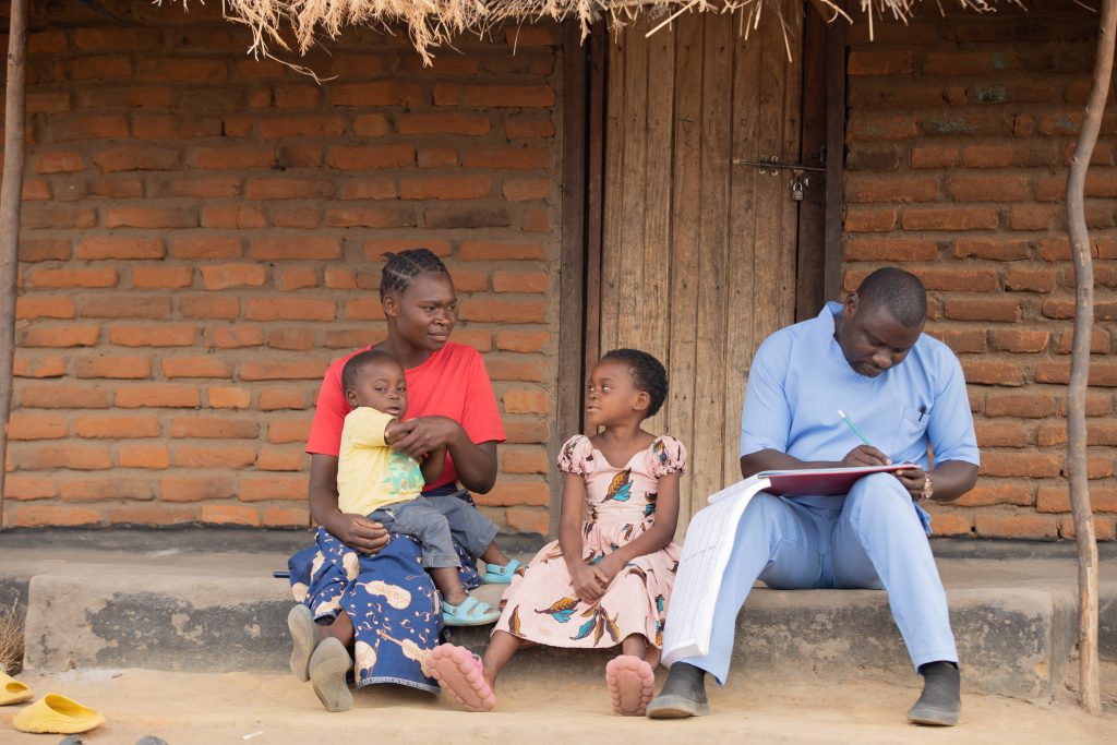 A mother sits outside on a step with her young son on her lap and her daughter beside her. To their right, a health worker in blue scrubs and a large notebook sits, speaking to the mother.