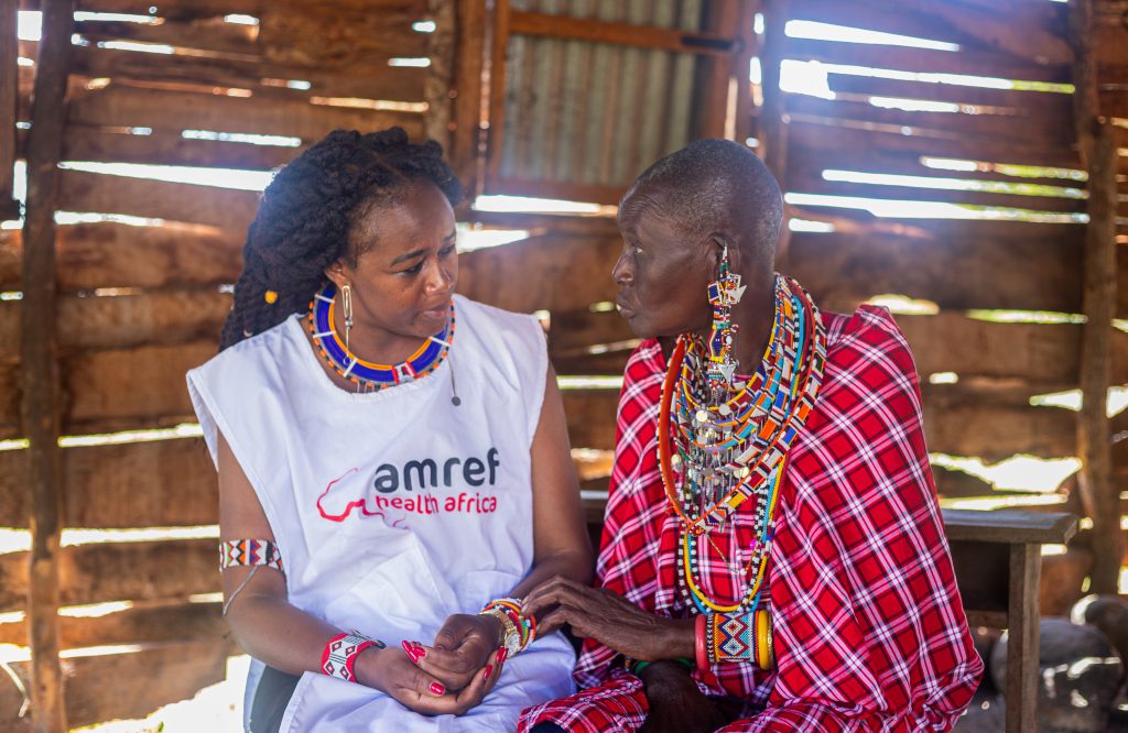 Cynthia and an older woman sit beside each other talking. Cynthia wears an Amref tabard and the woman is in tradional Maasi red dress with lots of beaded jewelry.