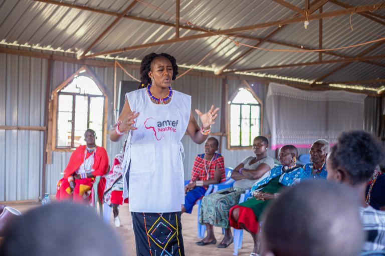 Cynthia stands in the middle of a circle of people listening to her. She is speaking passionately. She wears an Amref tabard.