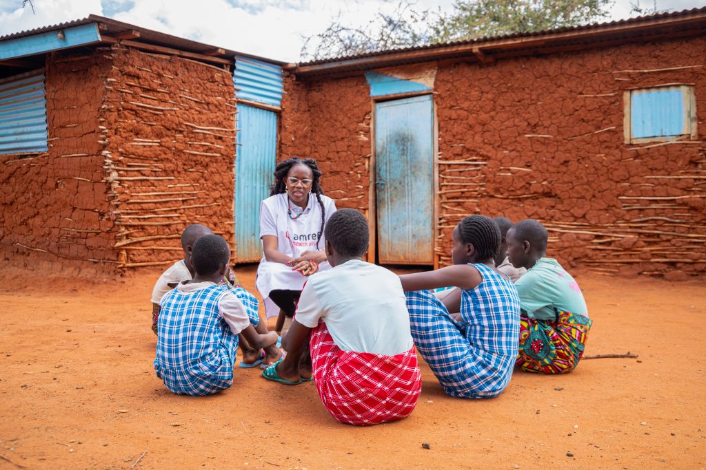 Cynthia sits on a low stool. six girls sit on the ground listening to her. Cynthia wears an Amref tabard and the girls are in in blue or red checked dress.