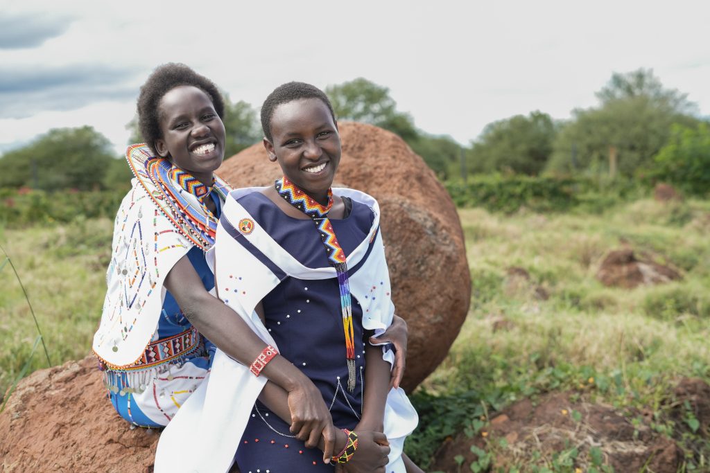 Jane Nkotai, End FGM/C advocate sits to the left, behind her sister. Both are wearing traditional dress and smiling (c) Amref Health Africa/Wesley Koskei