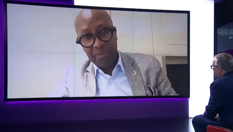 Still of Dr Githinji in his office on a screen in the channel 4 news studio. Matt Frei sits to the right engaging him in coversatioon.
