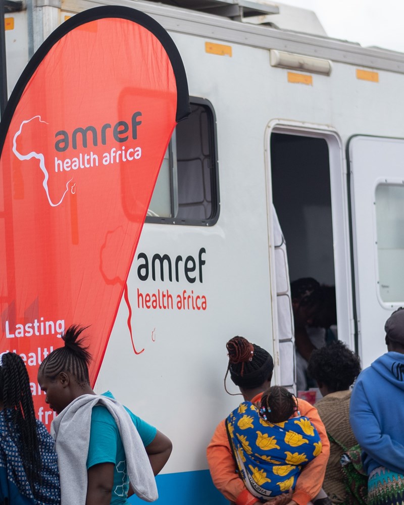 An Amref labelled mobile clinic, with a red teardrop banner with Amref written on it. People are waiting outside to be seen.