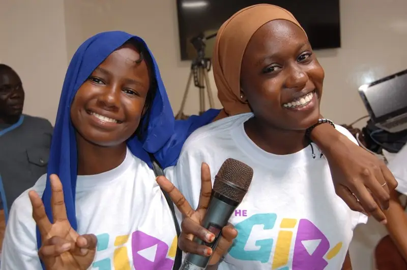 Two young women wear white T-shirts that have 'The Girl Generation' written on them. One wears a blue headscarf, the other brown and holds a microphone. They are smiling.