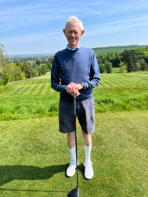 Fundraiser John Pool, a retiree stands holding a golf club, on the green. He's dressed in blue.