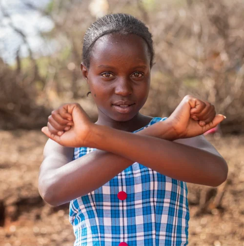 13-year-old Phoebe stands outside with her arms in front of her in a cross shape to represent 'no to FGM/C'. She wears a blue checked dress.