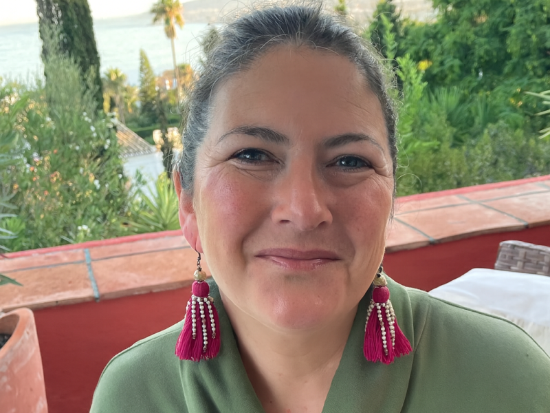 Headshot of Madeline. She smiles outside in a green top and long pink earrings.